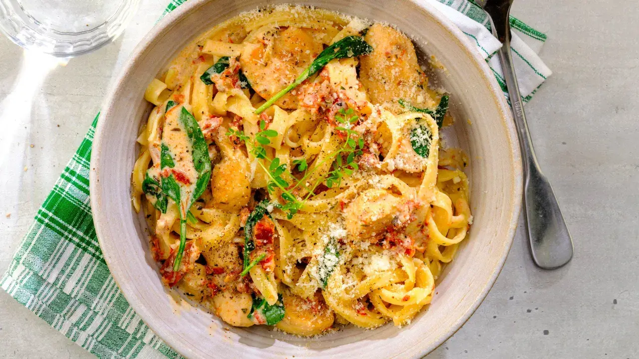 En skål med krämig pasta med soltorkade tomater, kyckling och spenat, toppad med riven parmesan och färska örter.