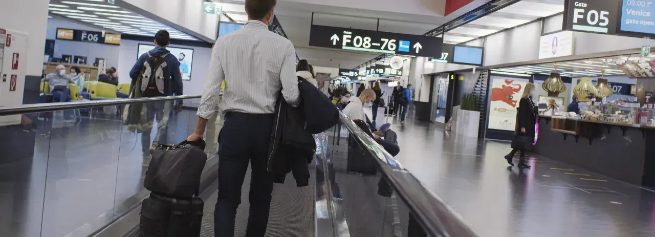 Resenärer på rullband på flygplatsen, skyltar visar vägen till gate F08-76. En man med resväskor på väg mot centrum.