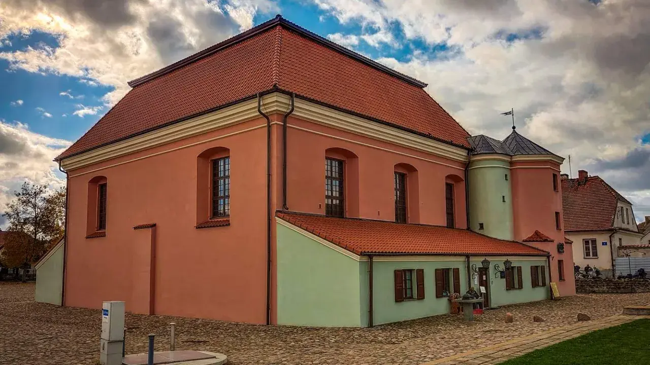 Tykocin co zobaczyć: zabytkowa Wielka Synagoga z różową fasadą i zielonym aneksem, na brukowanym placu pod burzliwym niebem.