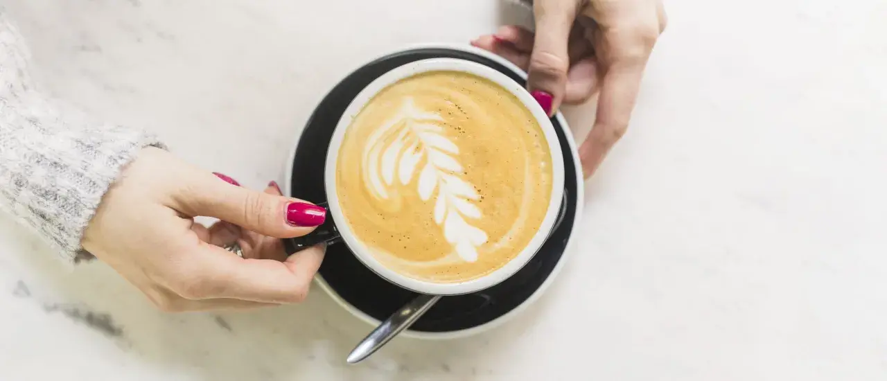 Une main tient une tasse de café latte avec un dessin de feuille. Un guide pour comment faire un latte parfait.