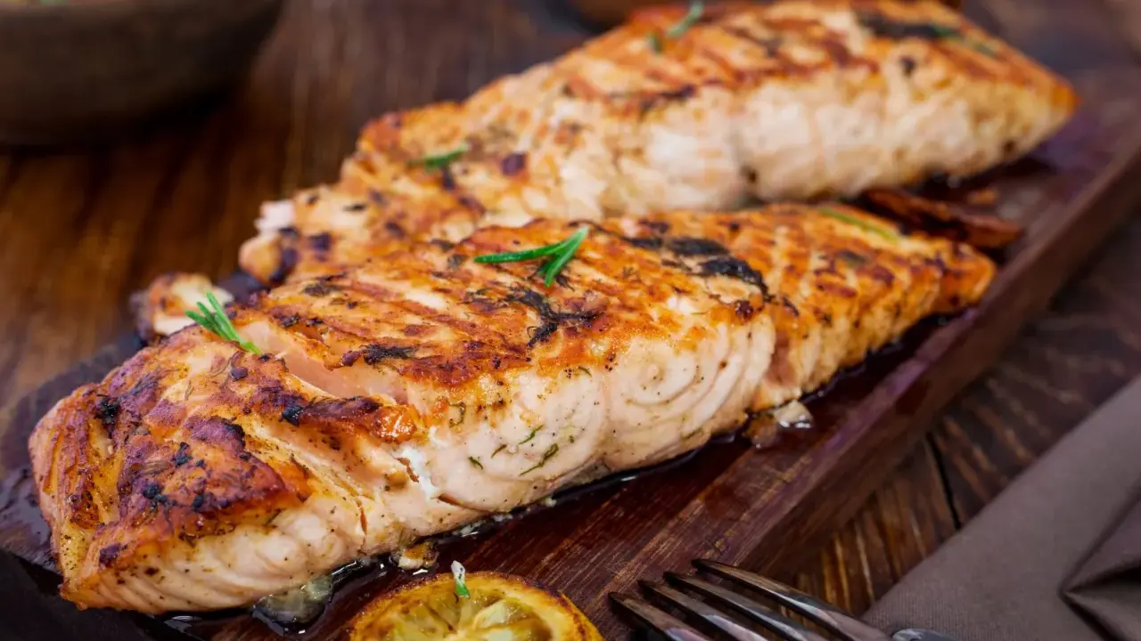 Två saftiga laxfiléer, perfekt grillade på halster, ligger på en träbricka. En citronskiva och rosmarinkvistar pryder fisken.