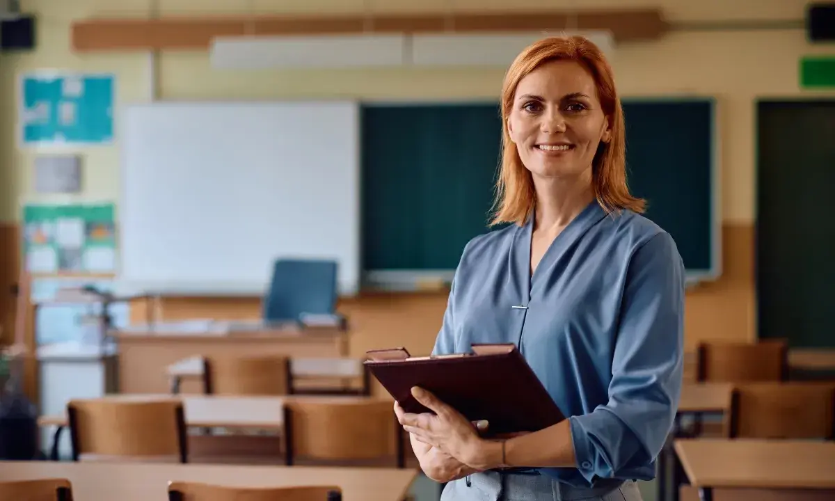 Uśmiechnięta nauczycielka z rudymi włosami trzyma teczkę. W tle klasa z ławkami i tablicą. Posiada legitymację nauczyciela.