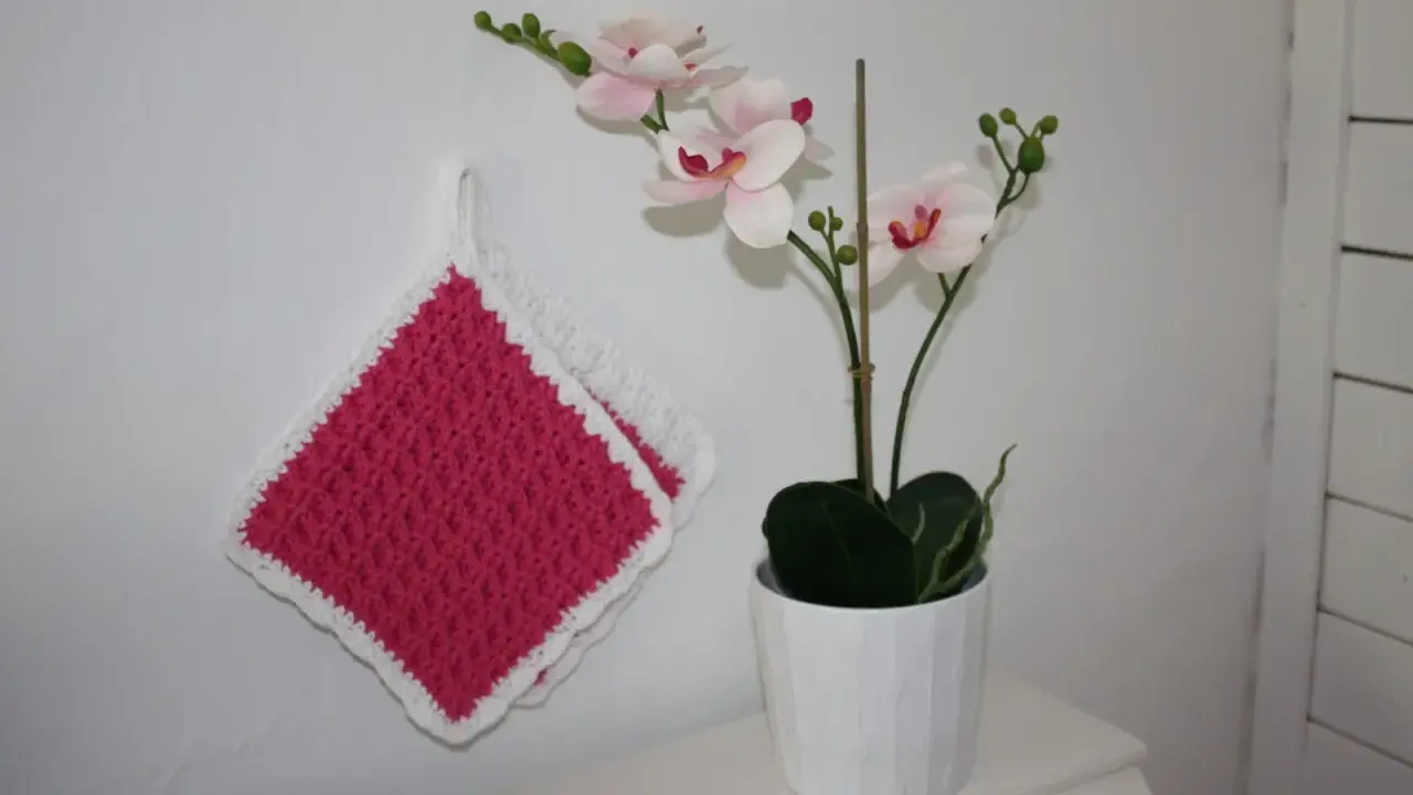 Två tjocka, virkade grytlappar i rosa med vit kant hänger på en vit vägg bredvid en orkidé i kruka.