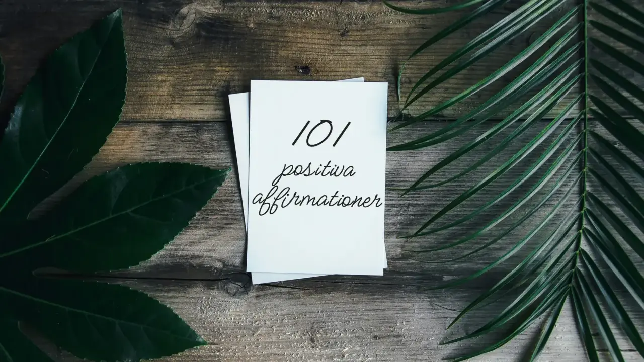 En hög med papper med texten "101 positiva affirmationer" på en träbakgrund, omgiven av gröna blad. En vacker påminnelse om affirmation svenska.