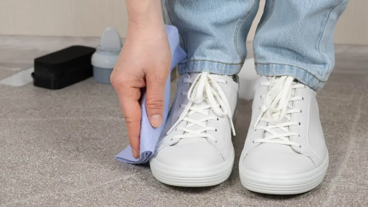 Persona limpiando zapatillas blancas con un paño azul. A su lado, un cepillo y un bote de limpiador.