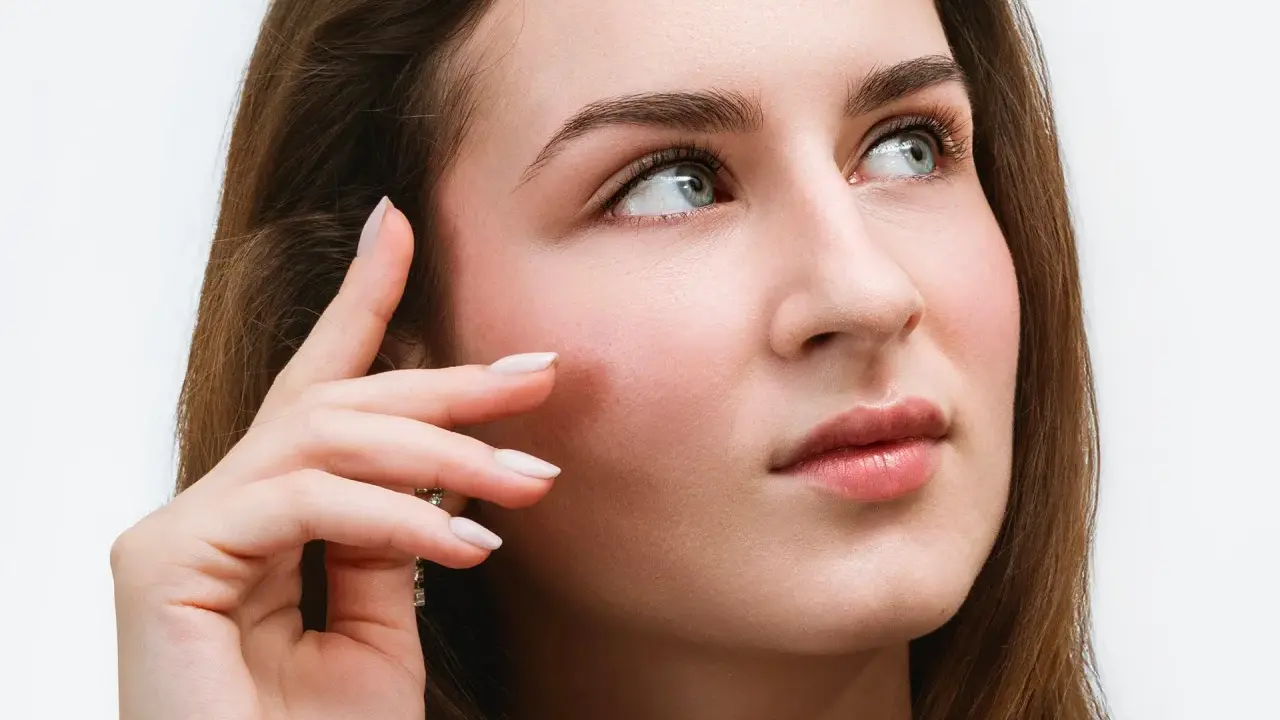 Une jeune femme aux yeux bleus regarde au loin, sa main effleurant sa joue. Ses cils, sublimés par un **tatouage cils**, accentuent son regard captivant.