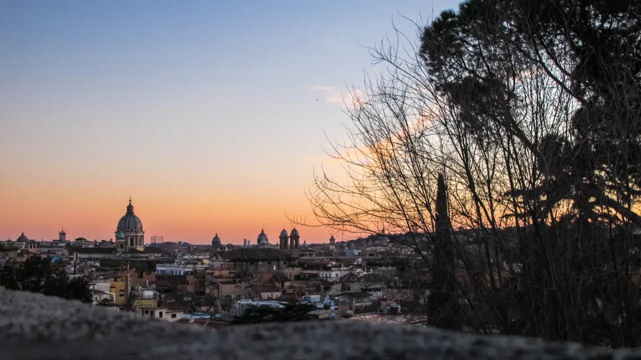 Un magico pincio roma tramonto con cupole che si stagliano contro il cielo aranciato, alberi spogli in primo piano.