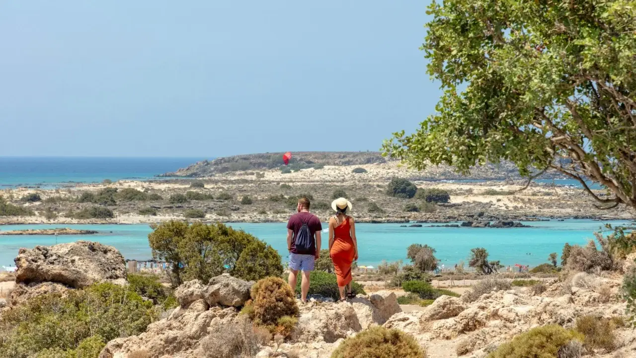 Ett par njuter av utsikten över Elafonissi, Kreta, med dess turkosa vatten och steniga landskap. En röd fallskärm syns i fjärran.