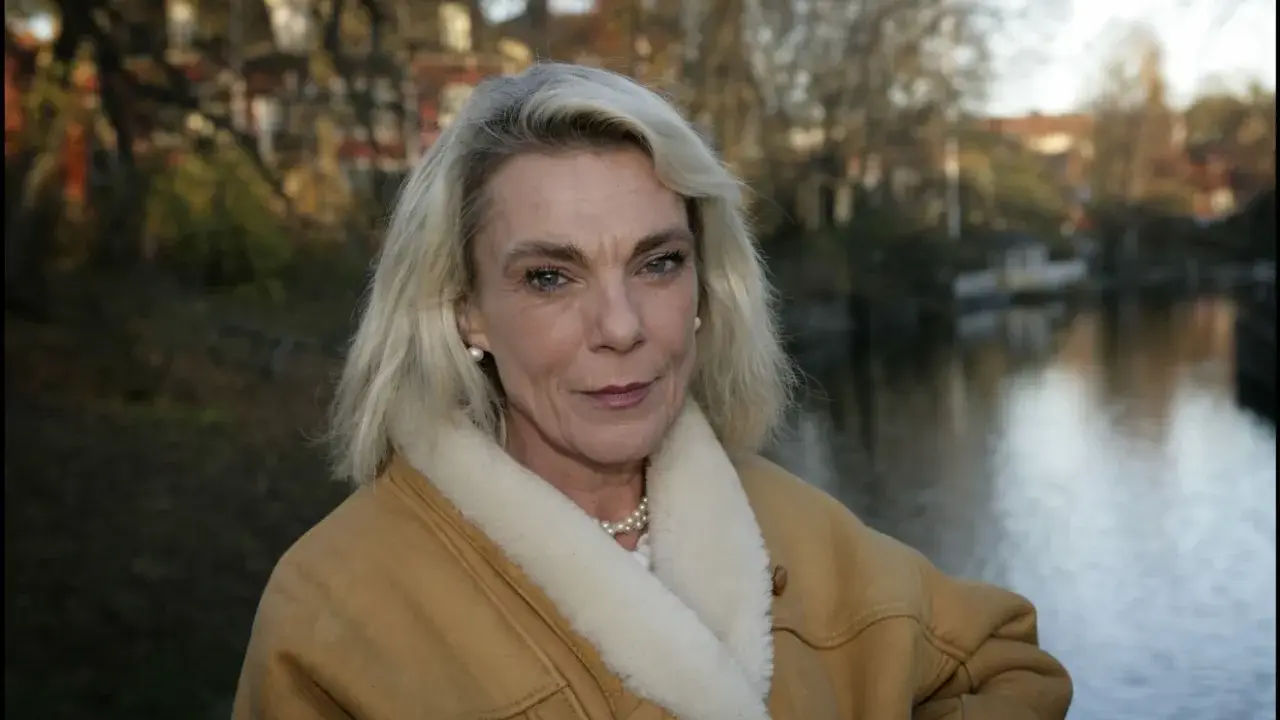 Pia Degermark idag, med sitt blonda hår och en varm fårskinnsjacka, står vid en vattensamling med träd och hus i bakgrunden.