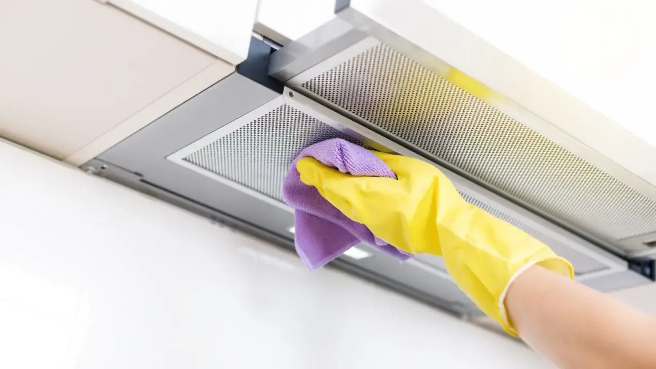 Mano con guante amarillo y paño morado limpiando la campana extractora por dentro.