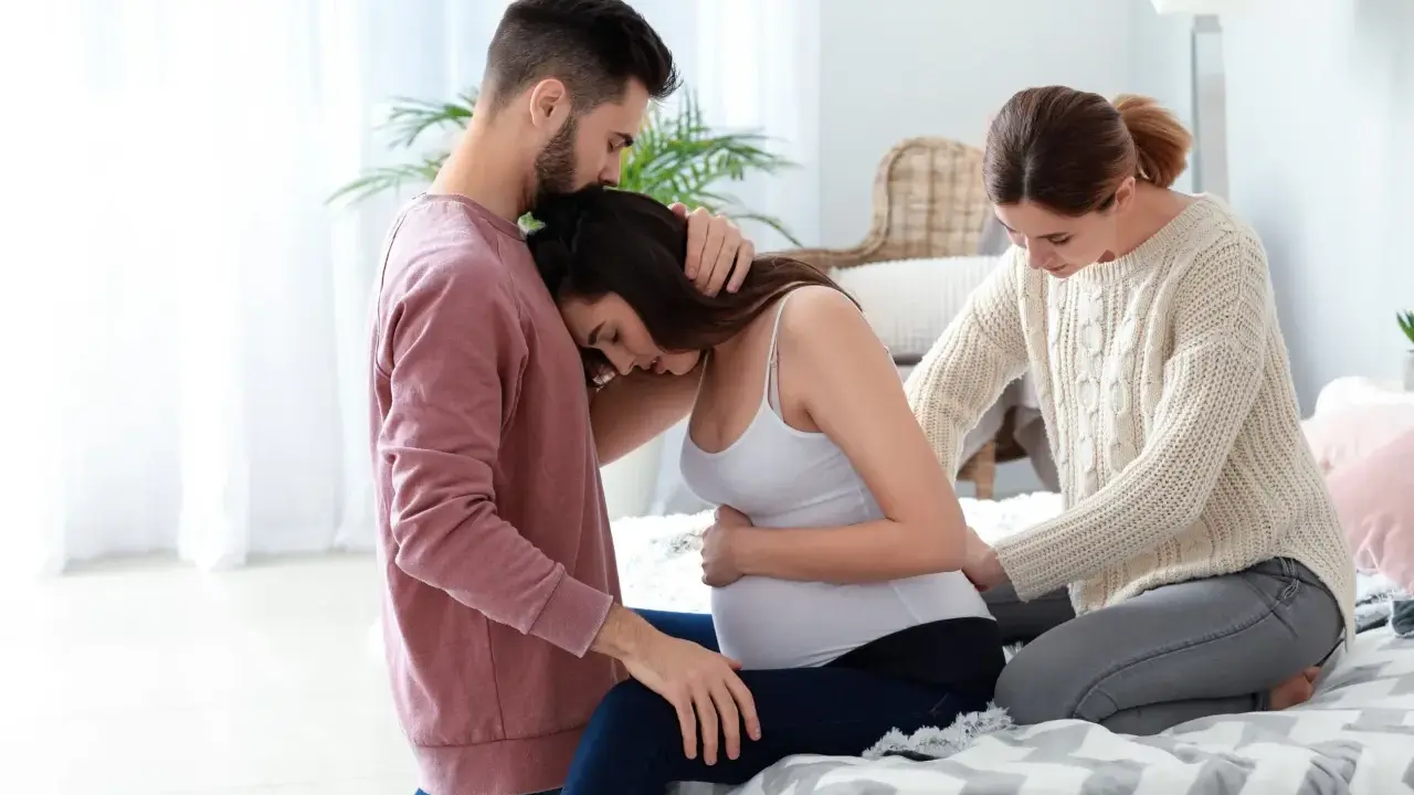 Mężczyzna obejmuje ciężarną kobietę, która opiera głowę na jego ramieniu. Obok siedzi doula, która delikatnie dotyka jej brzucha, oferując wsparcie.