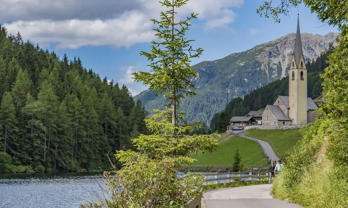 Passeggiata sul lago di Valdurna, con boschi rigogliosi, una chiesa alpina e montagne sullo sfondo.