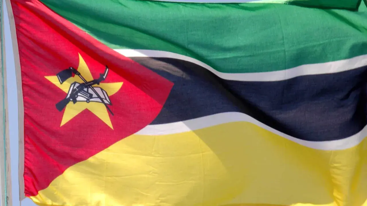 Flaga Mozambiku: zielono-czarno-białe pasy, czerwony trójkąt z żółtą gwiazdą, karabin AK-47 i książka.