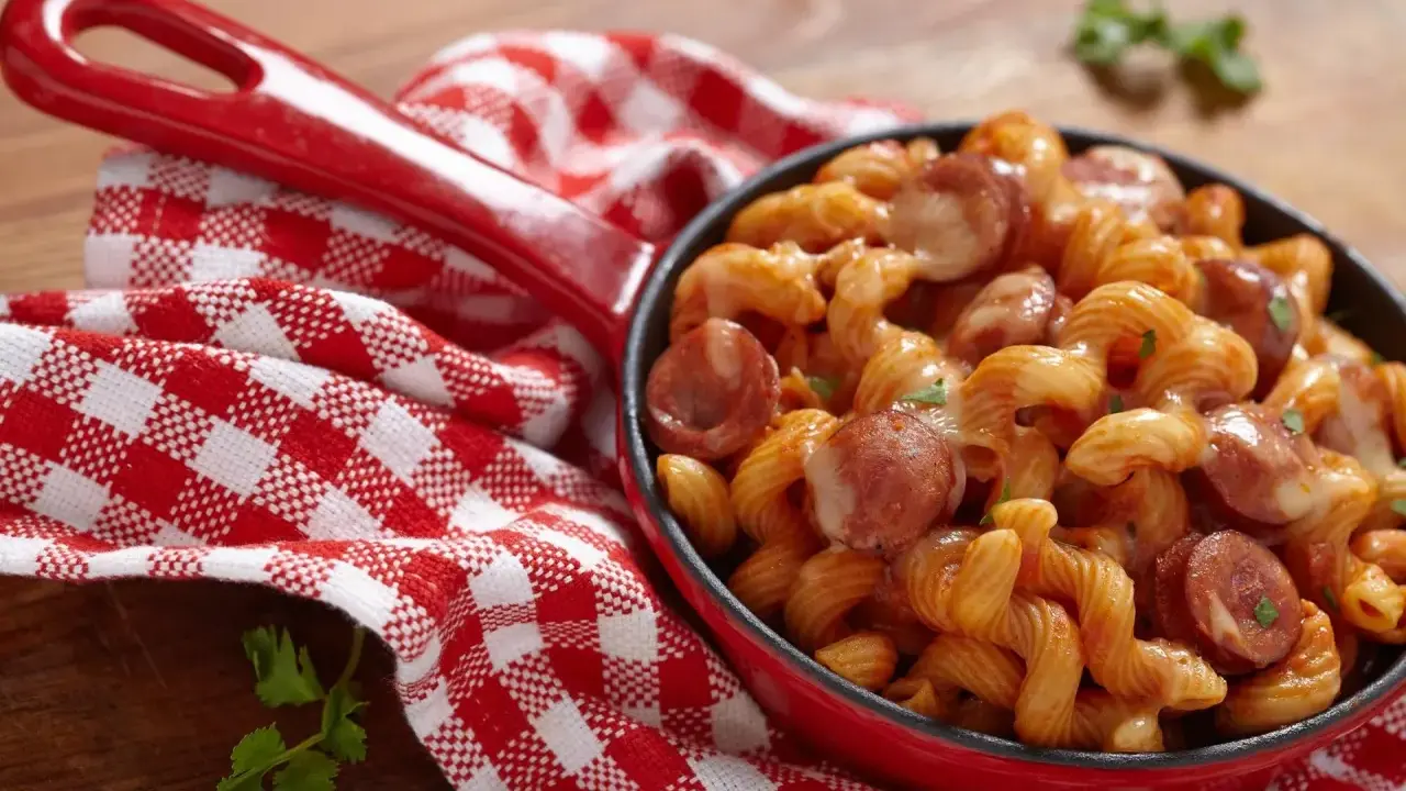 Pyszne spaghetti z kiełbasą w sosie pomidorowym, podane w czerwonej patelni, obok czerwono-białej kratkowanej serwetki.