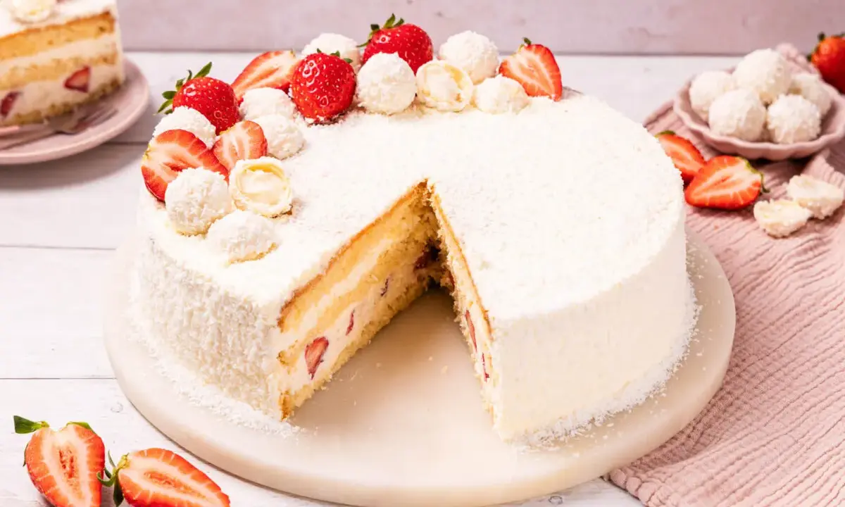 Eine köstliche Erdbeer Raffaello Torte, verziert mit frischen Erdbeeren und Kokosraspeln. Ein Stück ist bereits angeschnitten.