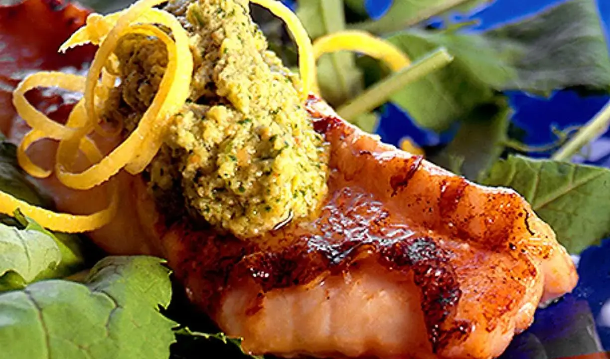 Grillad havsabborre med en grön topping och citronskal, serverad på en bädd av gröna blad.