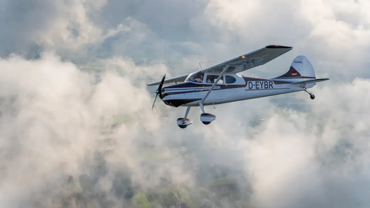 Eine braun-weiße Cessna 170 fliegt durch die Wolken über grünen Feldern.