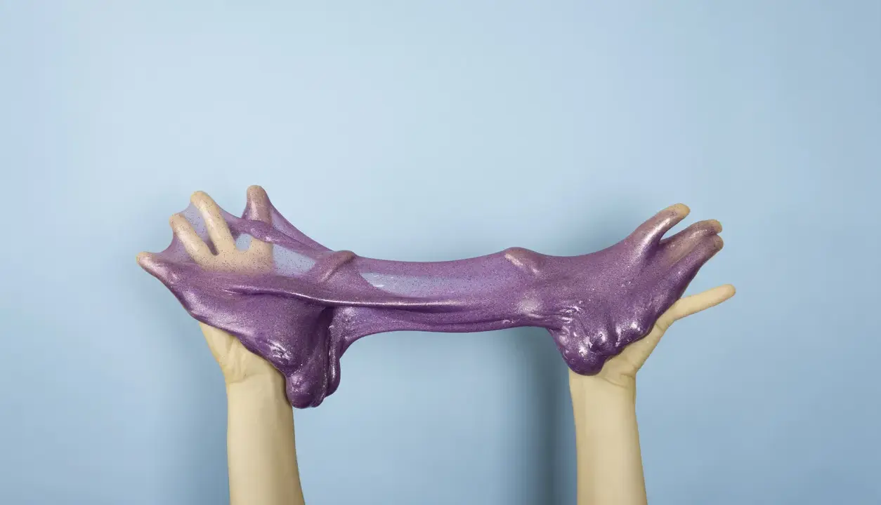 Manos estirando un brillante slime casero morado con purpurina sobre fondo azul.