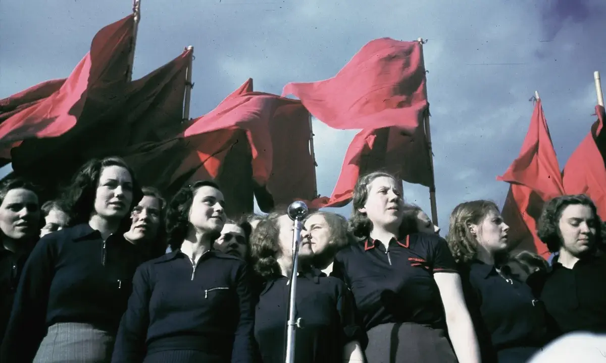 En grupp kvinnor deltar i ett 1 maj tåg, med röda fanor vajande i bakgrunden.