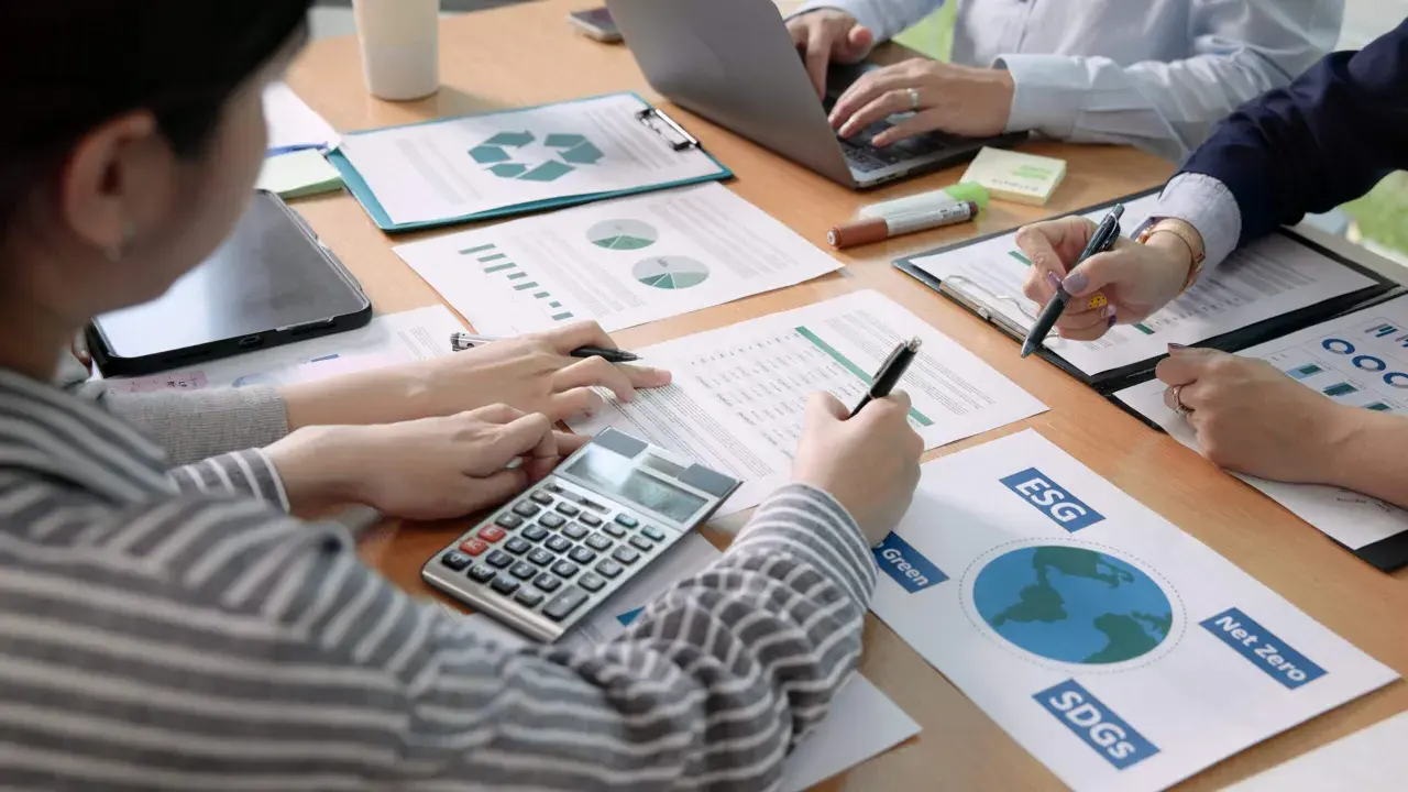 Des mains travaillent sur des documents financiers, une calculatrice et un ordinateur portable. L'ergonomie organisationnelle est au cœur de cette réunion axée sur le développement durable.