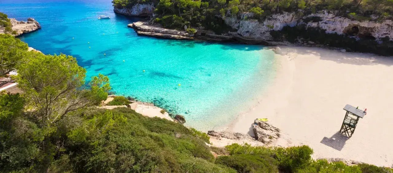 Traumhafte mallorca buchten mit türkisfarbenem Wasser, weißem Sand und grünen Pinien. Ein Boot ankert im Meer, eine Rettungsschwimmerturmbefindet sich am Strand.