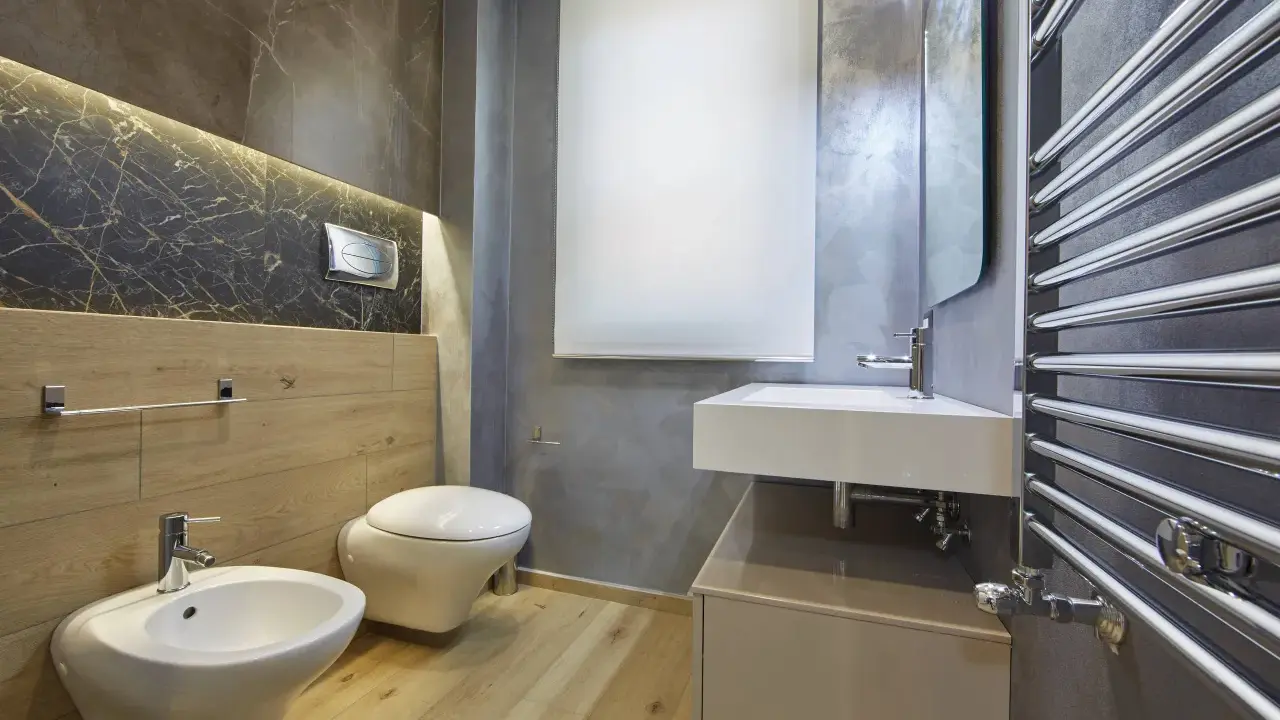 Bagno moderno con bidet, WC sospeso, lavabo e scaldasalviette cromato. L'illuminazione a LED sotto il marmo crea un'atmosfera elegante, rispettando l'altezza minima soffitto.