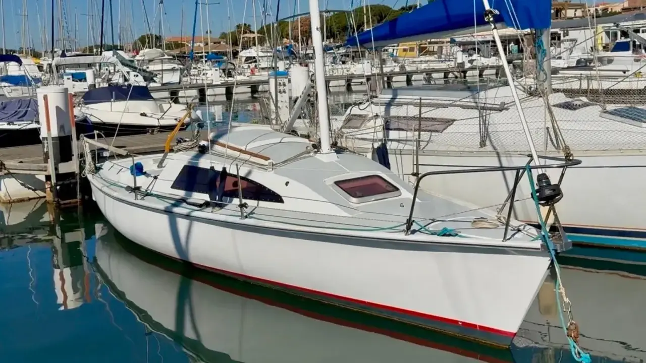 Un voilier blanc avec une voile bleue dans un port, reflété dans l'eau calme.
