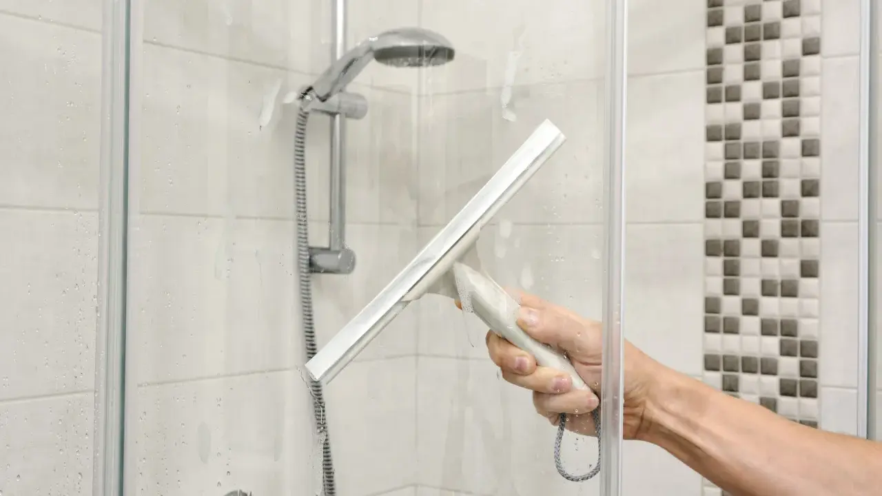 Hand mit Abzieher reinigt Duschkabine. Wasserflecken und Seifenschaum werden entfernt, um die Dusche zu putzen.