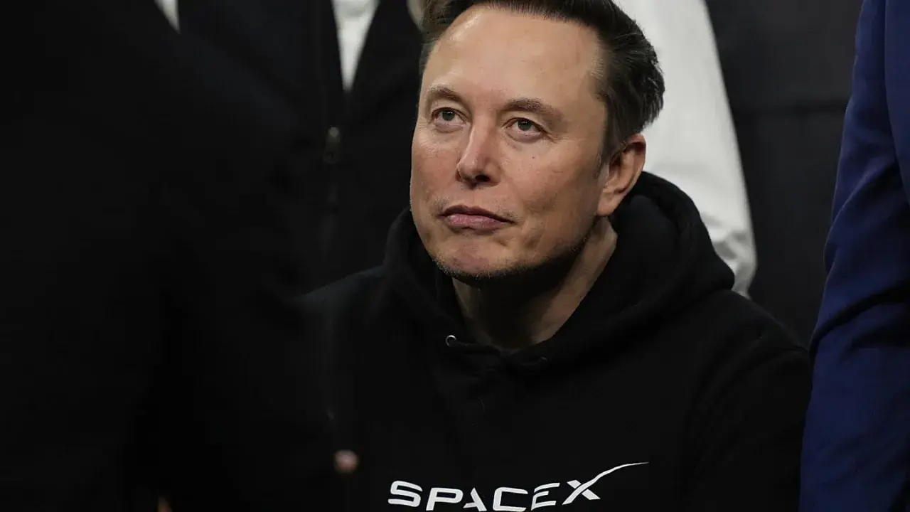 Elon Musk w bluzie SpaceX, zamyślony, w otoczeniu innych osób.