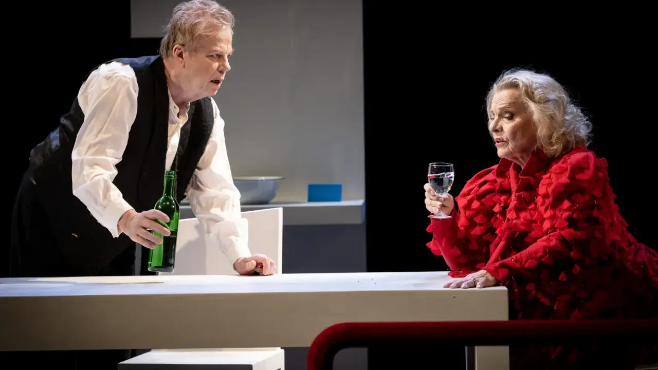 Krister Henriksson i en dramatisk scen. Han håller en grön flaska, medan en kvinna i rött med ett glas vatten ser på. Scenen påminner om hans roller i filmer och tv-program.