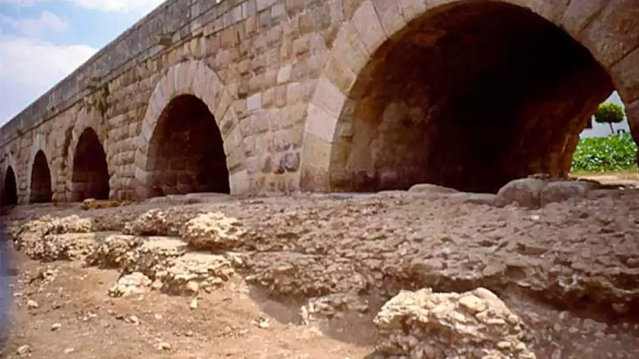 Antiguo puente de piedra con arcos, que es un puente de gran tamaño y robustez, con ruinas a sus pies.