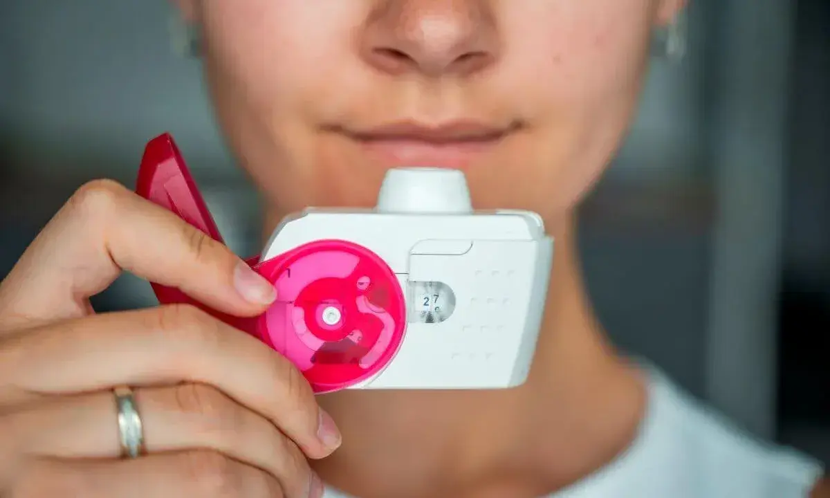 Nahaufnahme einer Person, die einen Pulverinhalator hält, bereit zur Anwendung. Der Inhalator ist weiß mit pinken Akzenten und zeigt die Dosis "27".