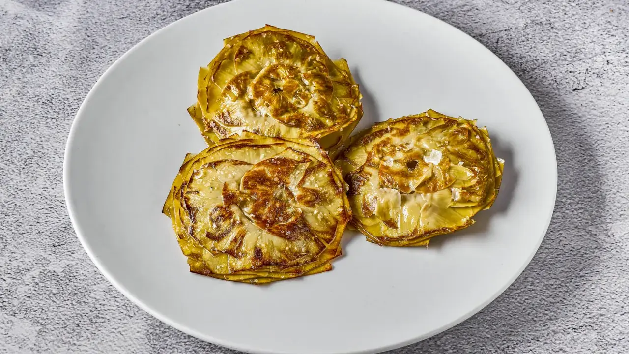 Tres alcachofas cortadas en rodajas, cocinadas a baja temperatura, con un toque de sal marina, servidas en un plato blanco.