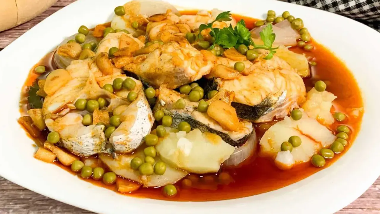 Merluza a la gallega con patatas, guisantes y ajo frito en una salsa rojiza.