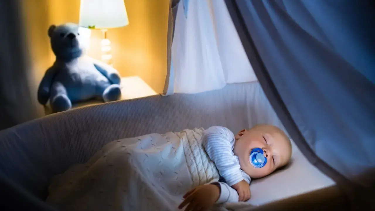 Bambino dorme sereno nella culla, con ciuccio, sotto una coperta calda. Un orsacchiotto veglia, simbolo di addormentamento autonomo.