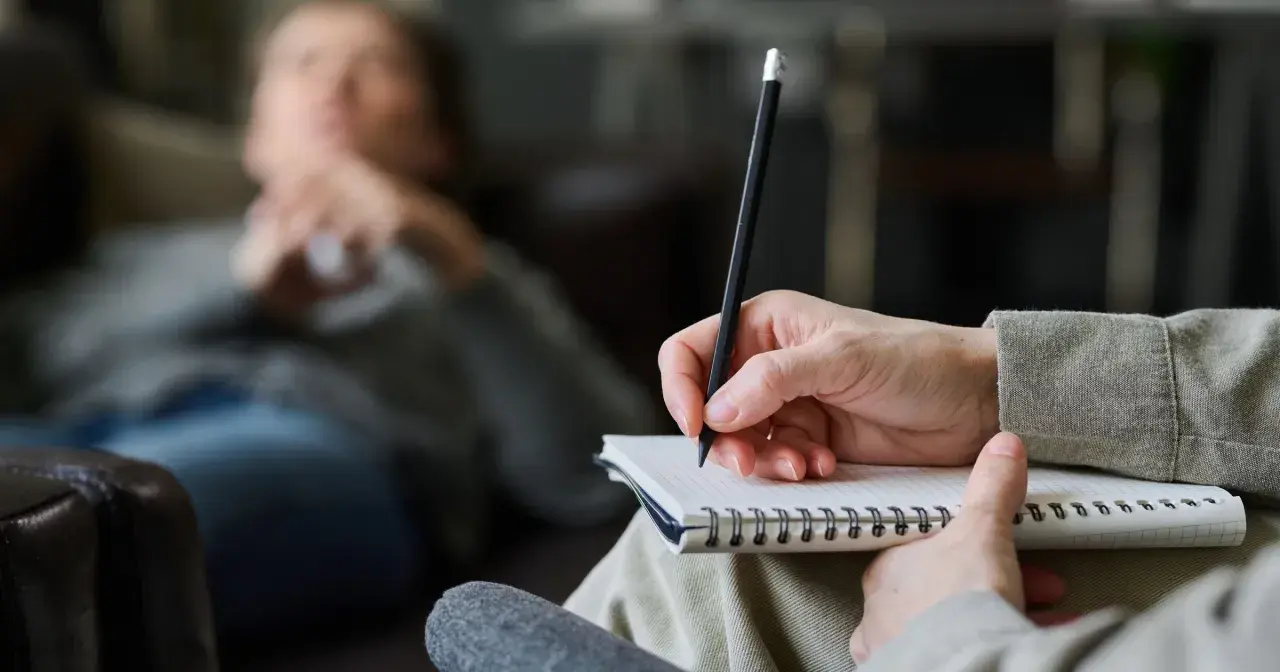 Psycholog pomaga zrozumieć siebie i swoje problemy. Na zdjęciu widać rękę terapeuty piszącą w notatniku, podczas gdy pacjent leży na kanapie.