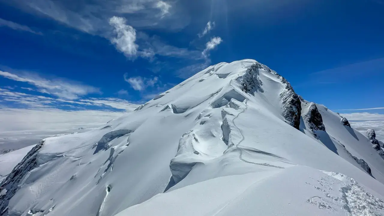 Śnieżna trasa na Mont Blanc, z widocznymi szczelinami lodowcowymi i śladami wspinaczy. Błękitne niebo z chmurami.
