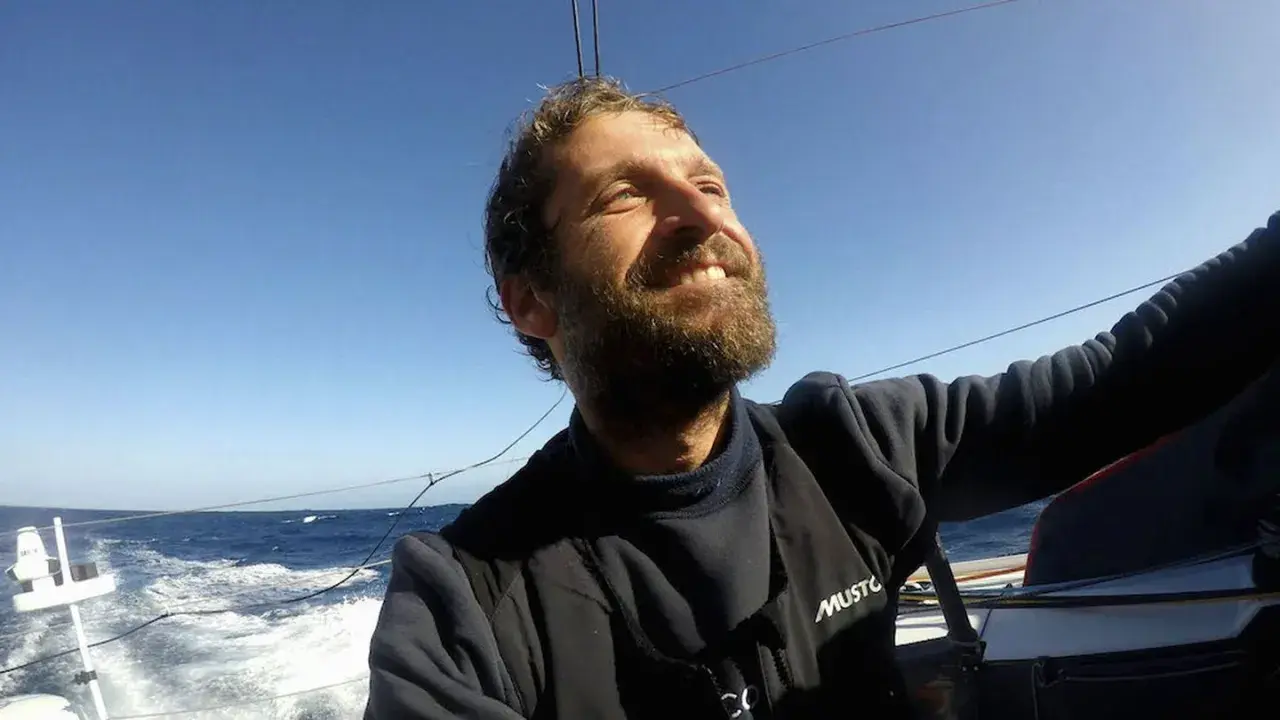 Un homme de mer souriant, le visage tourné vers le ciel bleu, navigue sur un voilier. L'océan agité et le soleil radieux témoignent de son aventure.