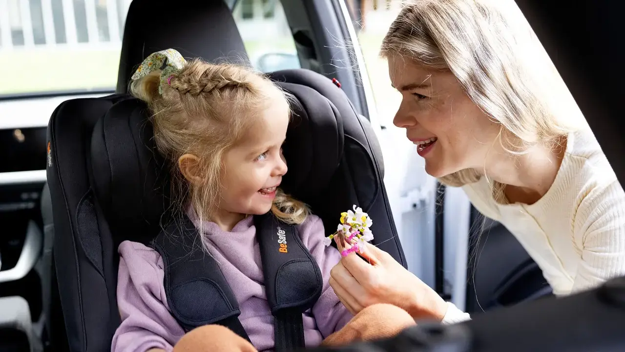 Uśmiechnięta mama daje córeczce w foteliku samochodowym bukiecik kwiatów. To miły test plus dla małej pasażerki.