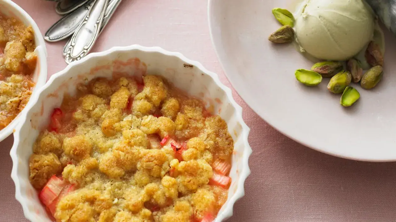 Rhabarber-Crumble in weißer Auflaufform, daneben eine Kugel Eis mit Pistazien. Ein köstliches Rhabarber Dessert.