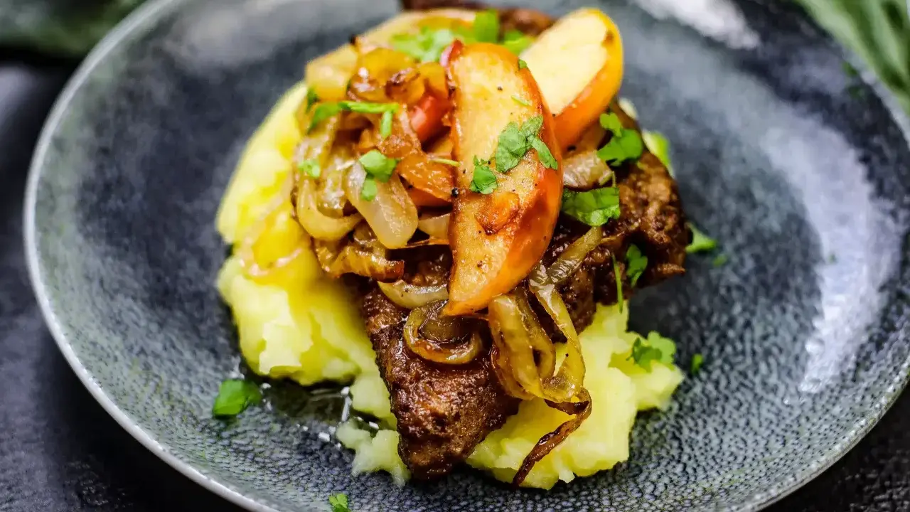Gedünstete Leber mit Apfelspalten, Zwiebeln und Kartoffelpüree auf einem dunklen Teller.