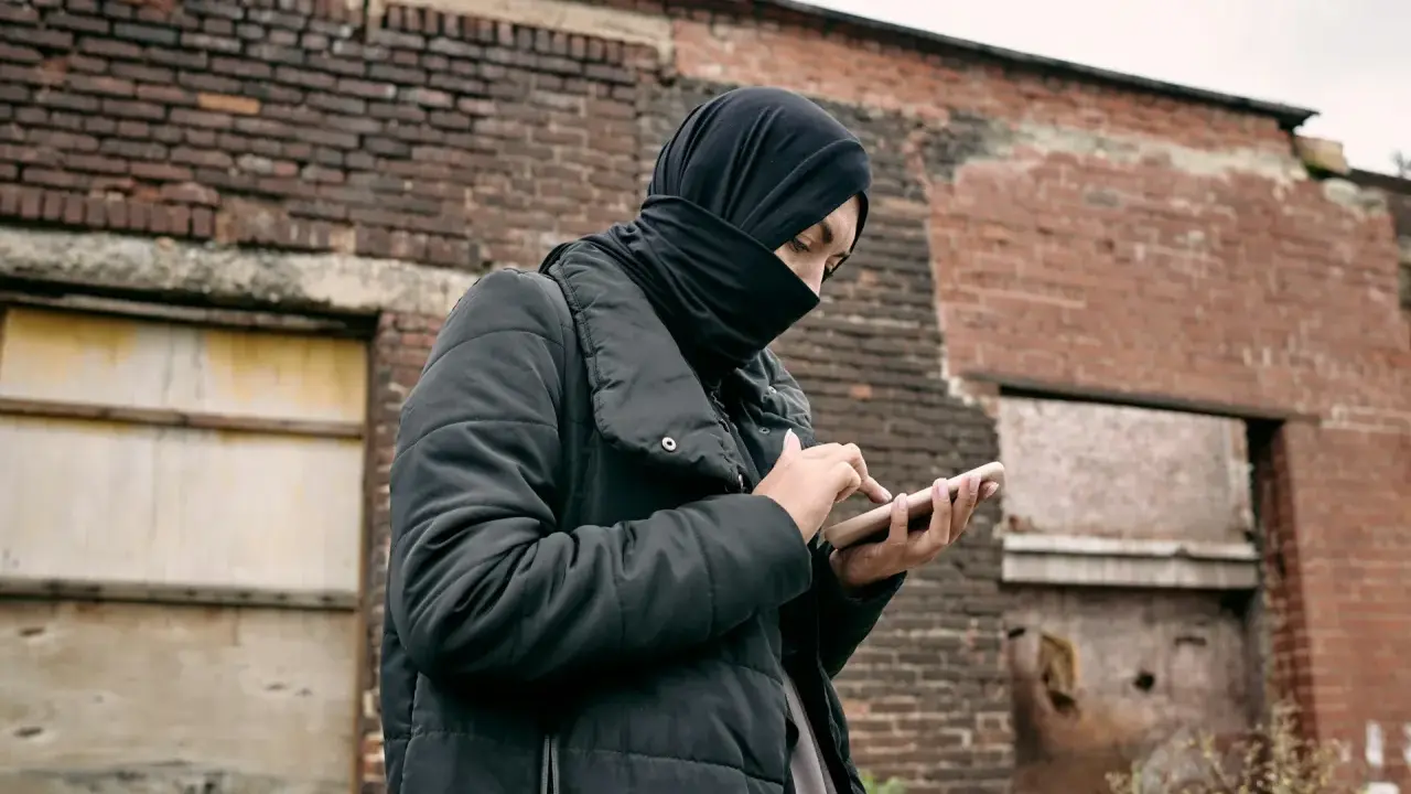 Osoba w czarnej chuście i kurtce trzyma telefon na tle zrujnowanej ceglanej ściany.