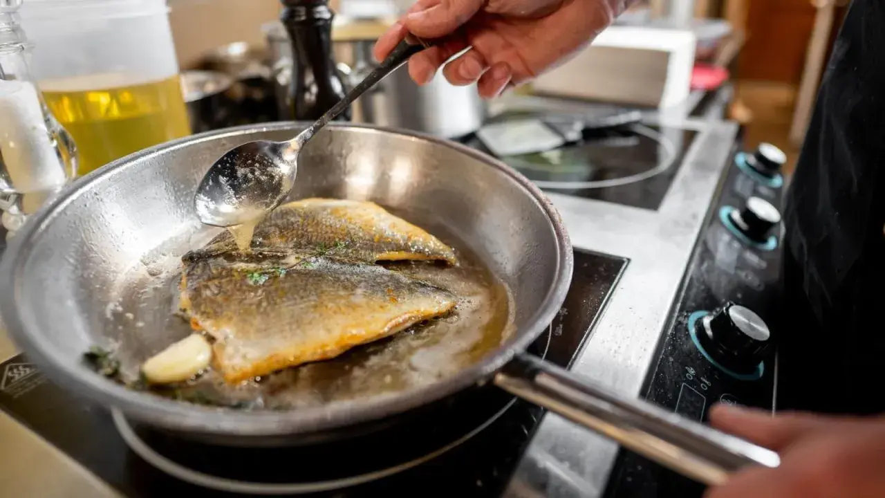 Un chef cocina pescado en una sartén de acero inoxidable, bañándolo con aceite caliente.