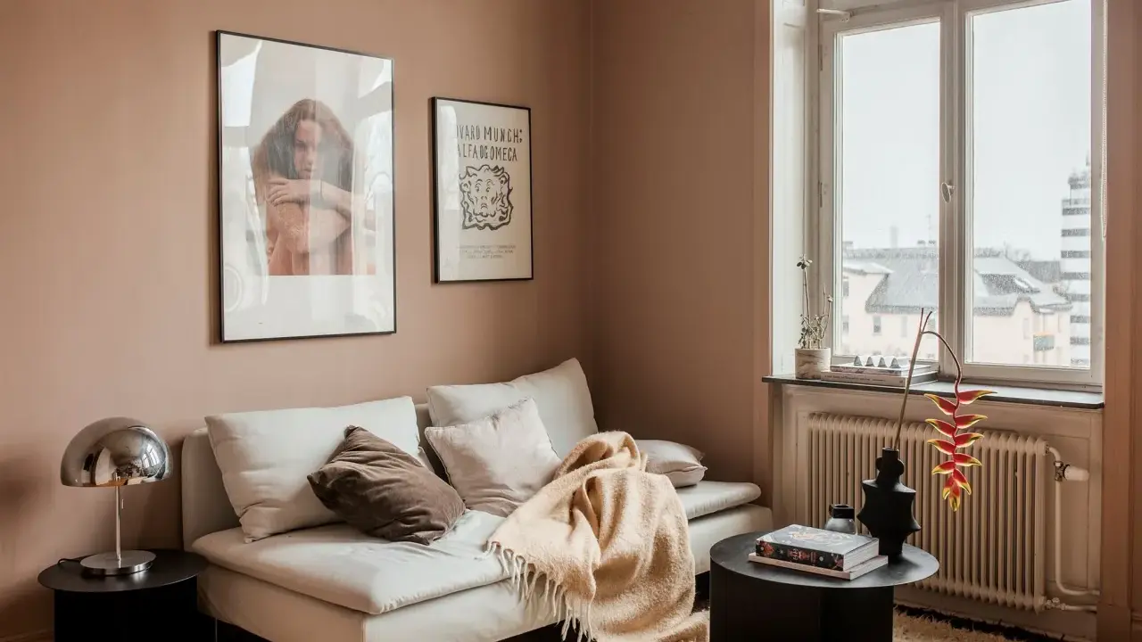 Un divano bianco accogliente con cuscini e una coperta color **rosa pastello** su un tappeto soffice. Alle pareti, due quadri e una finestra con vista.