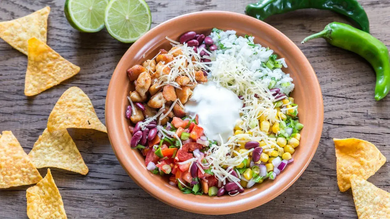 Bunter Taco Salat mit Hähnchen, Reis, Bohnen, Mais, Tomaten, Käse und Sauerrahm. Tortilla-Chips und Limetten daneben.