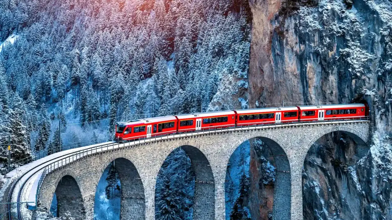 Un treno rosso attraversa un viadotto innevato, parte del suggestivo treno Bernina percorso, tra alberi coperti di neve e rocce imponenti.