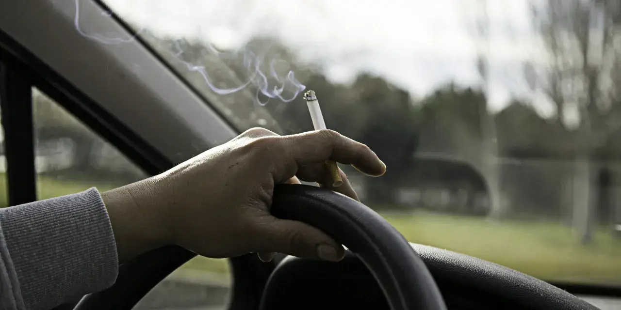 Mano sujetando un cigarrillo encendido sobre el volante. El humo se eleva mientras se puede fumar conduciendo.