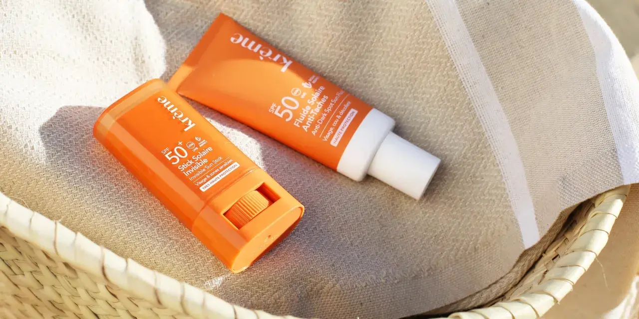 Deux produits solaires, un stick et un fluide, sont posés sur une serviette rayée. Le bouton du stick est visible, prêt à protéger du soleil.