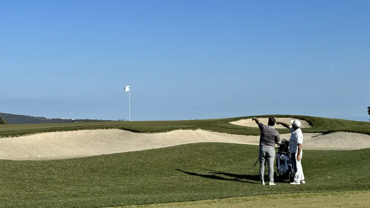 Zwei Golfer planen ihren nächsten Schlag auf dem Valderrama Golfplatz, mit einem Bunker und dem Fahnenmast im Hintergrund.