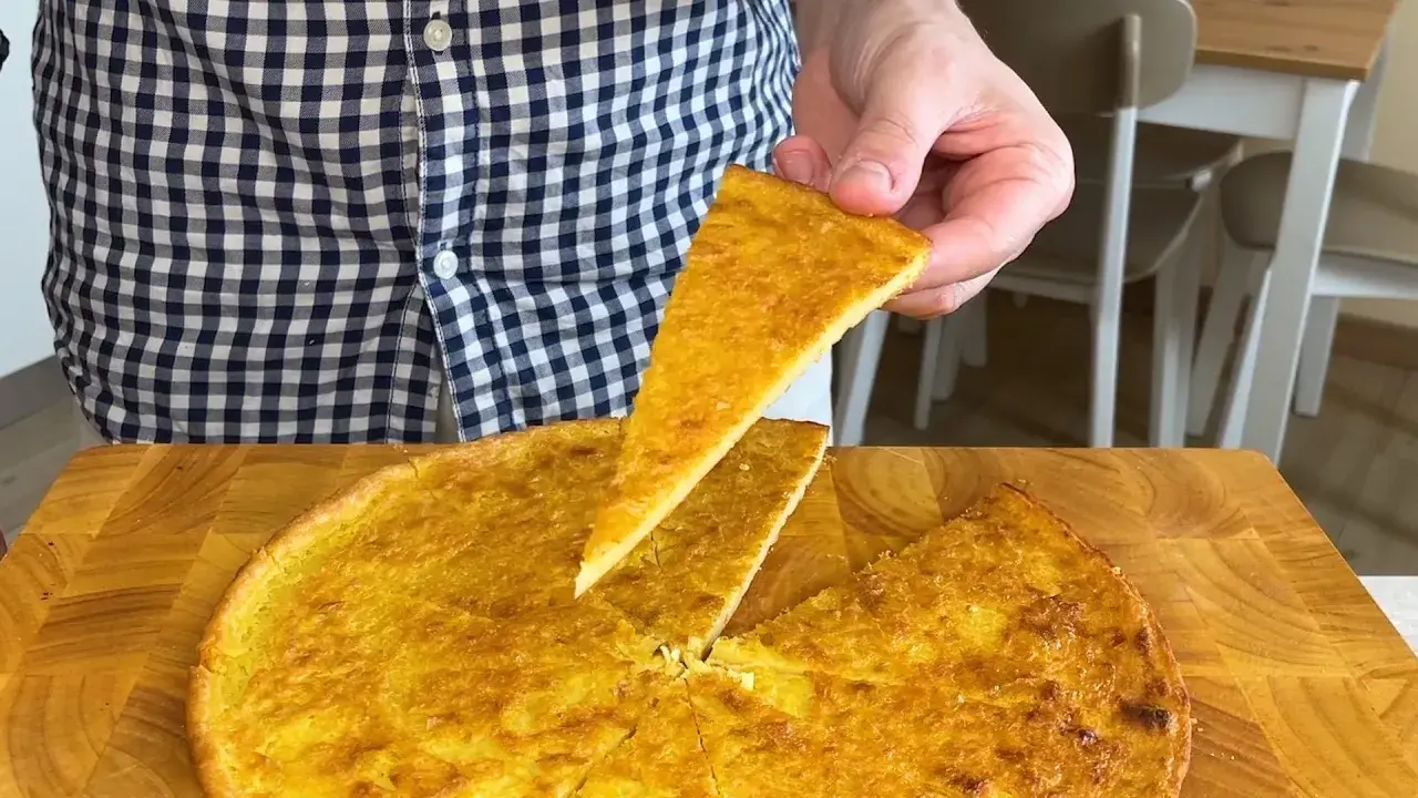 Una fetta di farinata di ceci dorata e croccante viene sollevata da una teglia rotonda su un tagliere di legno.